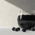 A minimalist glass bowl filled with glossy blueberry compote, surrounded by fresh blueberries on a clean white surface.