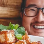 Best Ways to Cook Tofu: Smiling person holding a plate of crispy, golden-brown tofu garnished with sesame seeds and fresh cilantro.